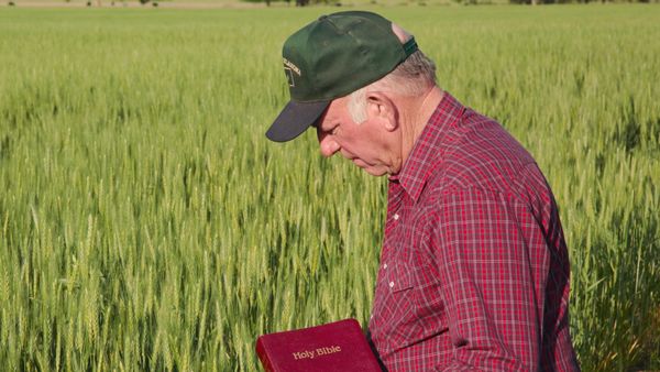 Photo of farmer in field where a flannel button-up and a green Oklahoma ball cap looking down and holding a red Holy Bible in his hand