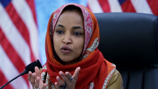 U.S. Rep. Ilhan Omar speaks into a micrphone and gestures with both hands, with American flags visible in the background. She wears a patterned orange and red hijab.