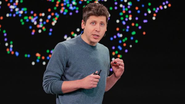 Image shows OpenAI CEO Sam Altman, a white man with brown hair, speaking in front of a dark backdrop. 