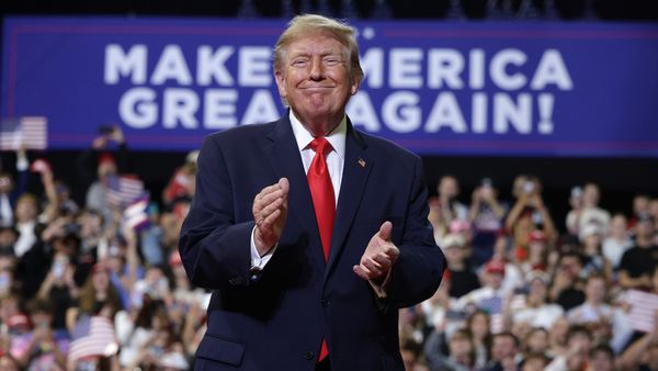 U.S. President Donald Trump, wearing a suit and red tie, claips his hands and smiles. In the background, a crowd of people and a "Make America Great Again" sign are visible.